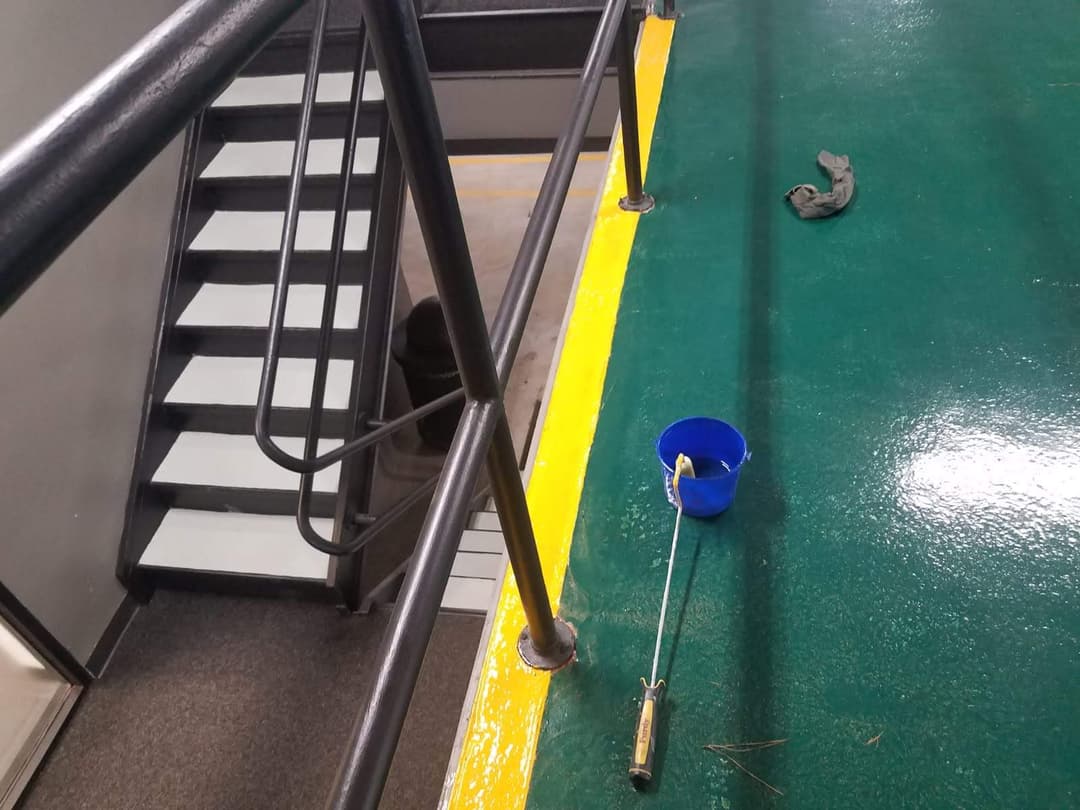 Staircase with black railings and green floor, blue bucket, and paint roller in workspace.