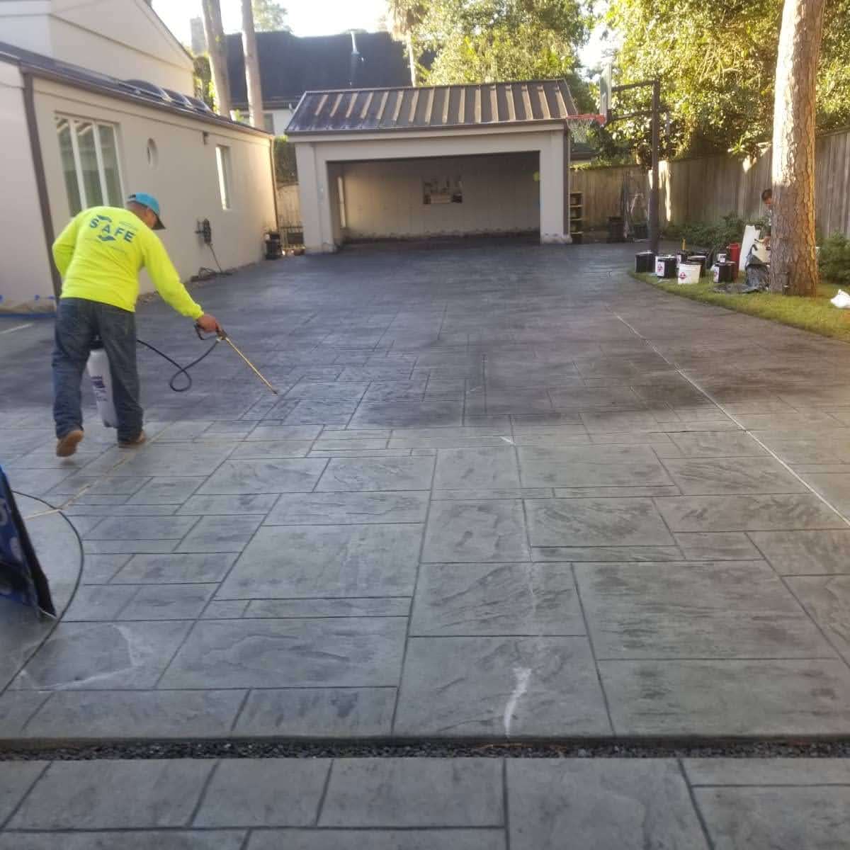 Worker applying sealant to a newly finished stamped concrete driveway in a residential yard.