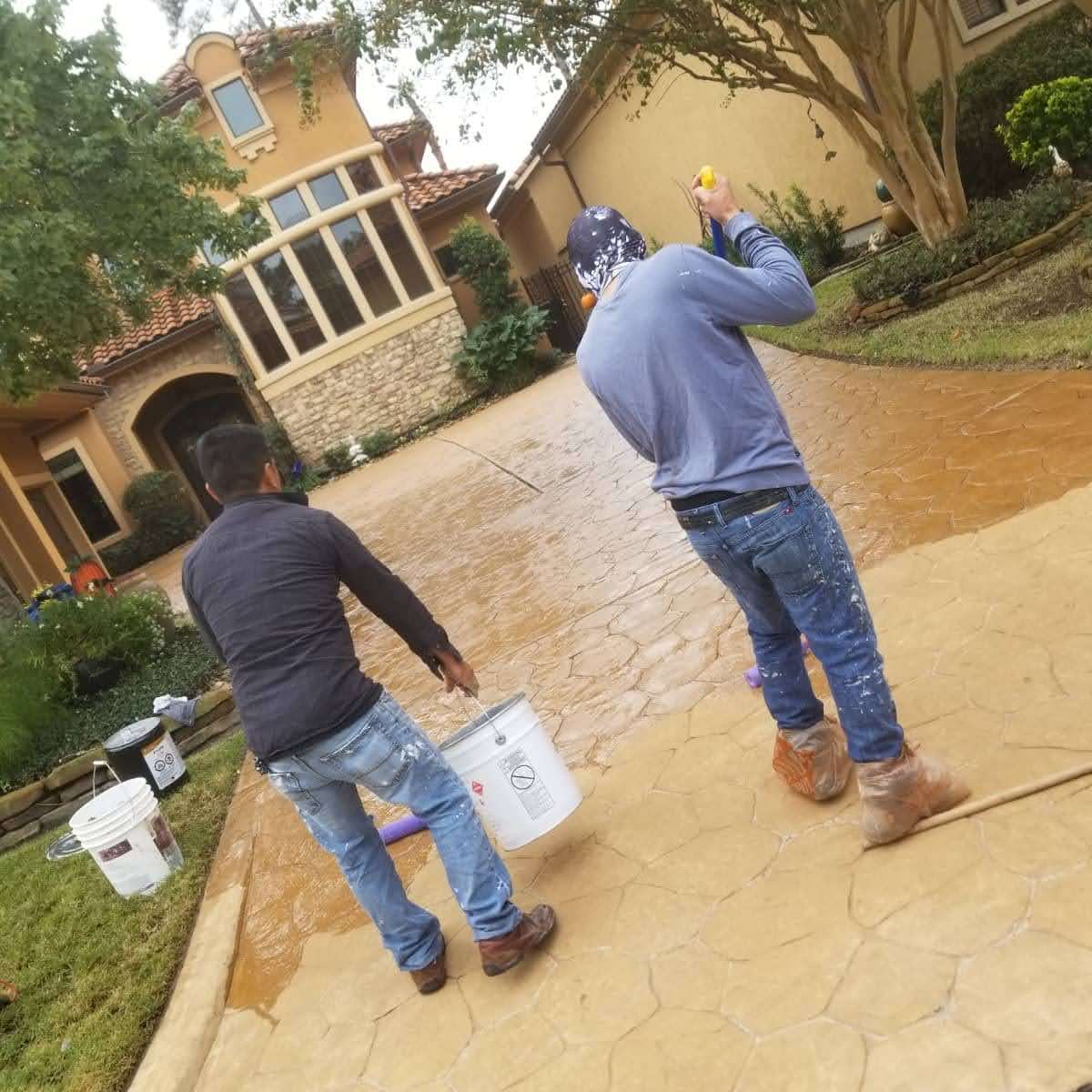 Workers applying decorative concrete finish on a residential driveway with tools and paint buckets.