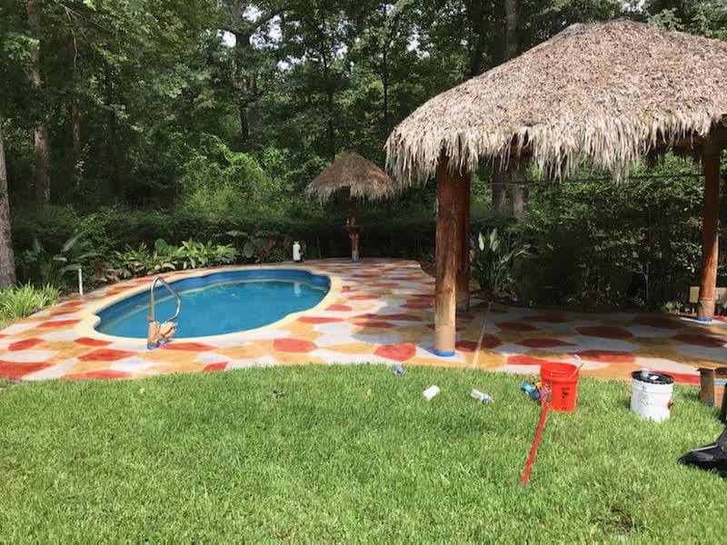 Tropical backyard with a pool, stone patio, and thatched-roof gazebo surrounded by lush greenery.
