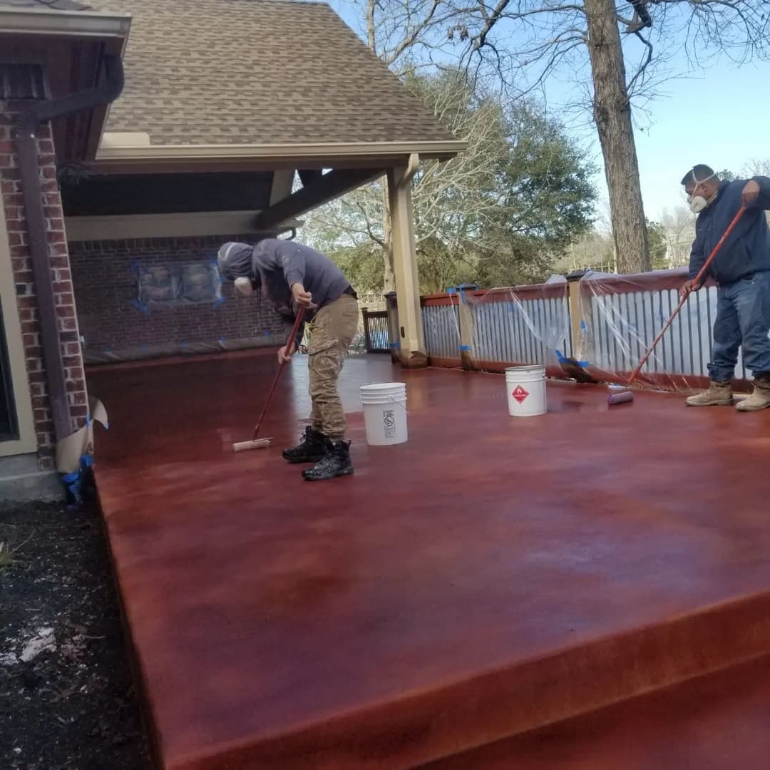Workers applying decorative concrete stain to a patio surface with rollers and buckets.
