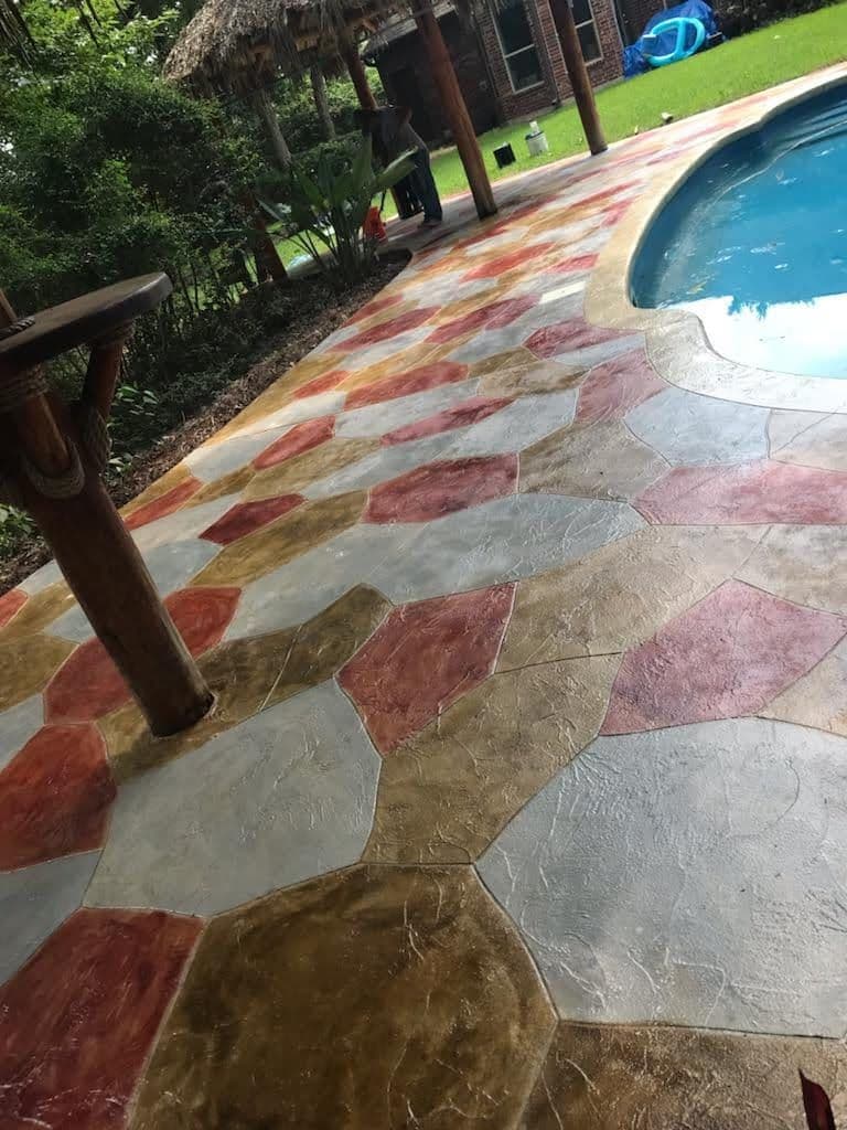Decorative stamped concrete patio around a pool with colorful stone patterns and lush greenery.