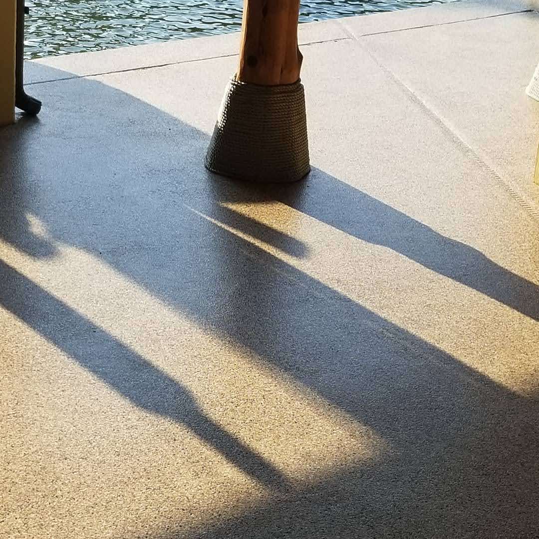 Shadow of a wooden post on a textured surface near water in soft sunlight.