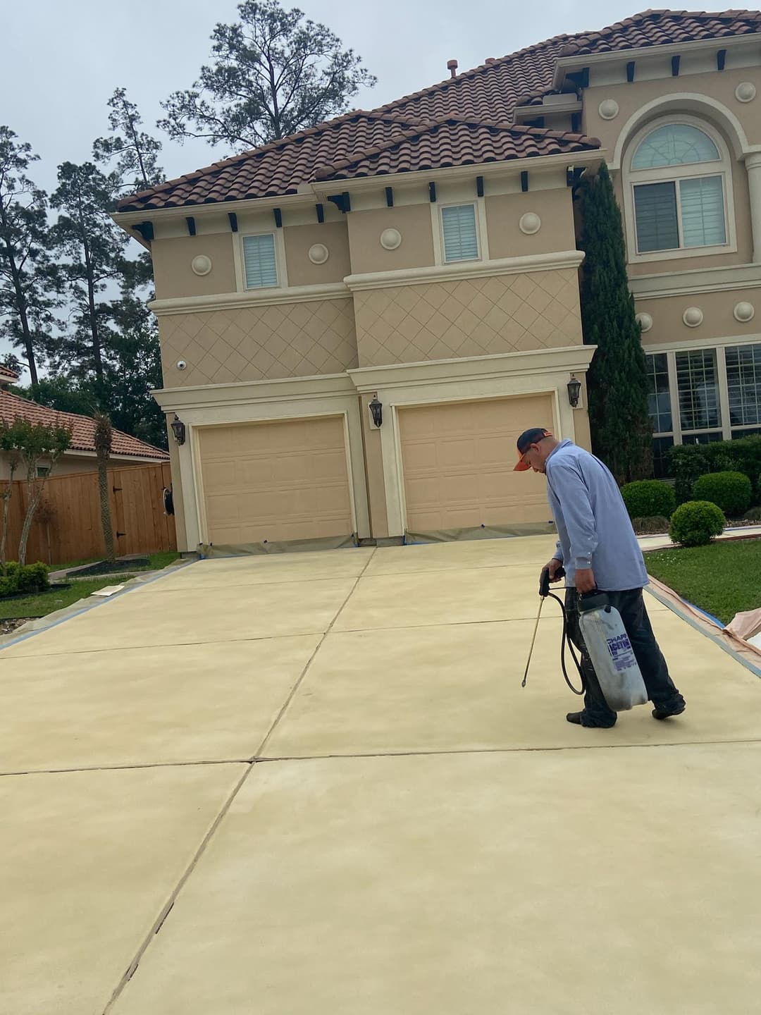 Worker applying sealant to freshly poured concrete driveway beside luxury home.