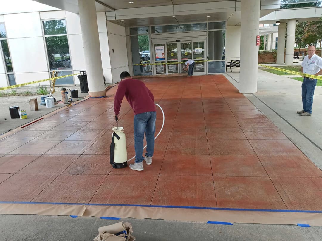 Worker applying sealant to freshly poured decorative concrete surface outside a building entrance.