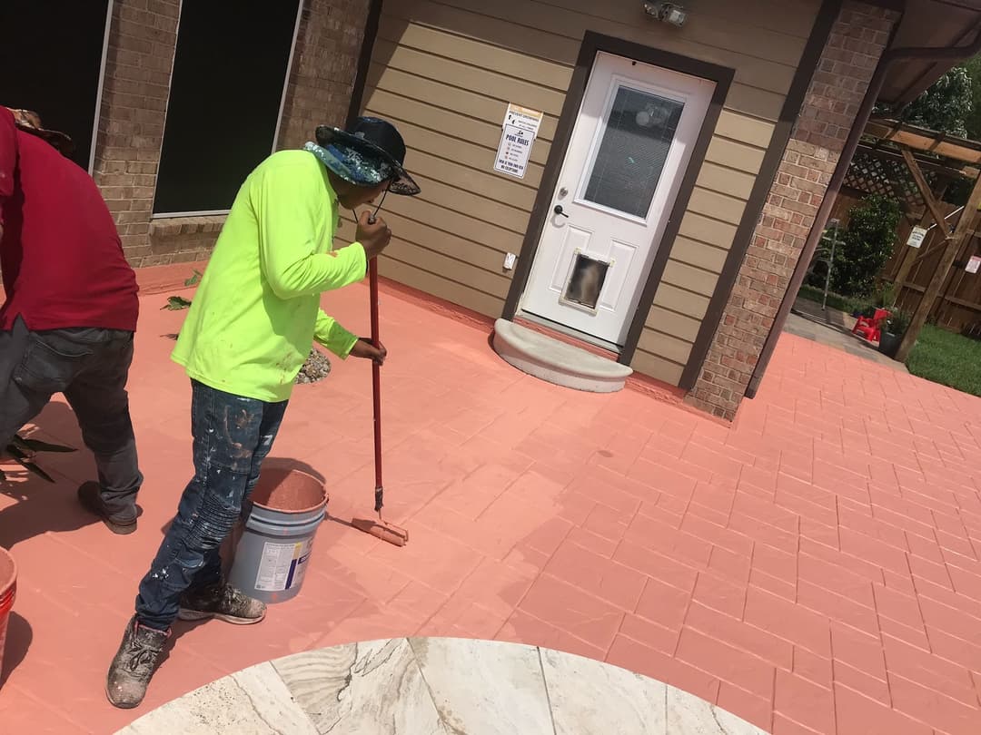 Worker applying sealant to a newly finished stamped concrete patio in bright sunlight.