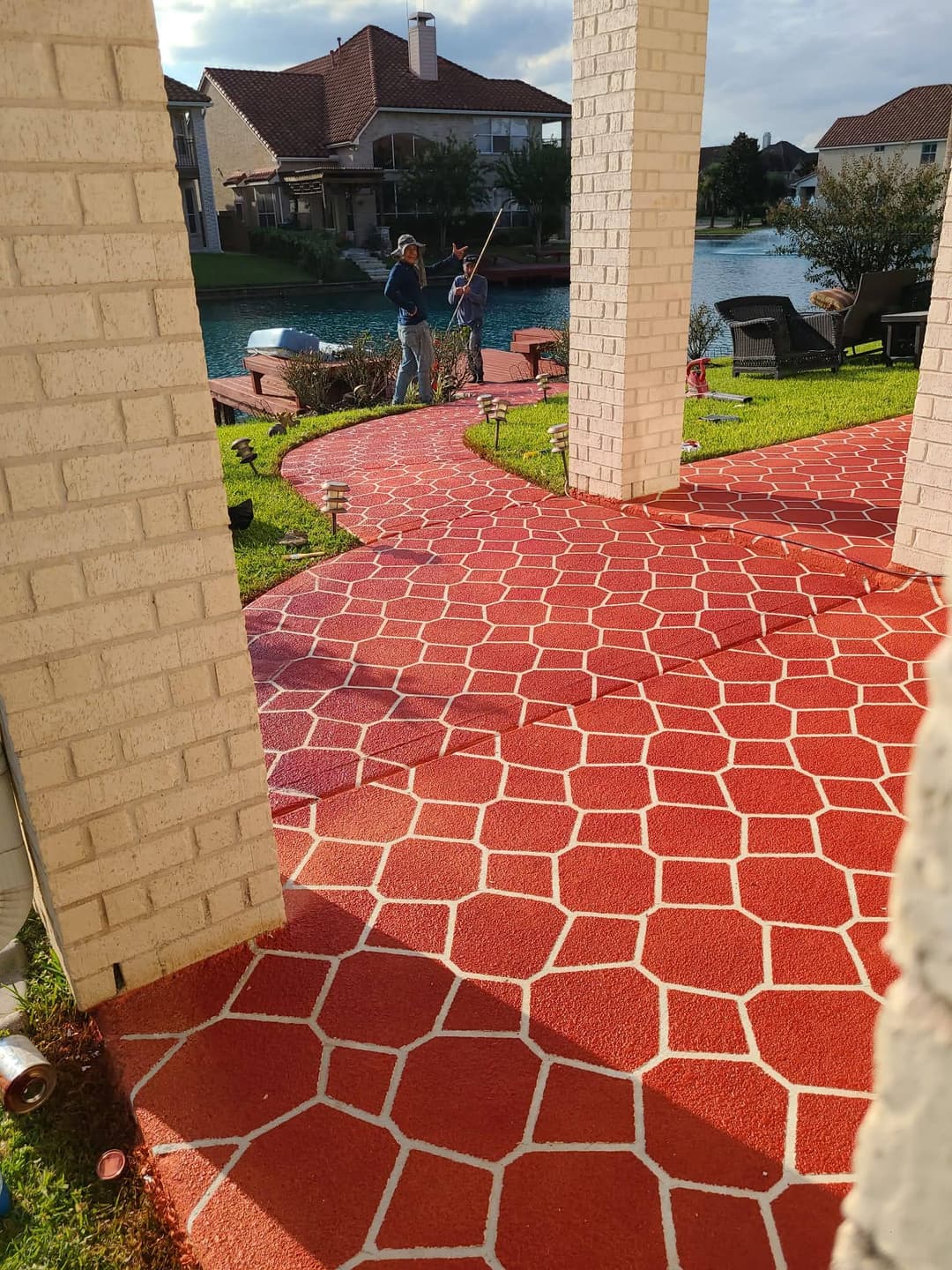 Men painting a red stone-patterned walkway near a serene lake and landscaped yard.