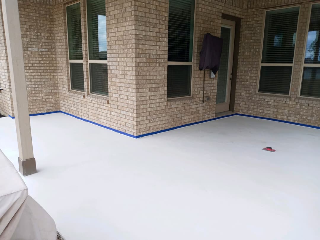 Freshly painted white patio floor with blue tape along edges, featuring brick wall and windows.