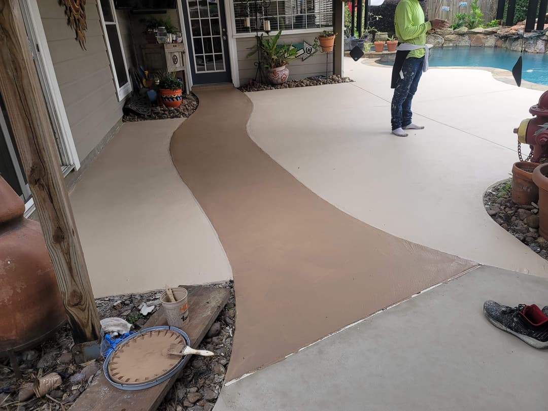 Person applying a brown coating on a patio, with potted plants and a swimming pool in background.