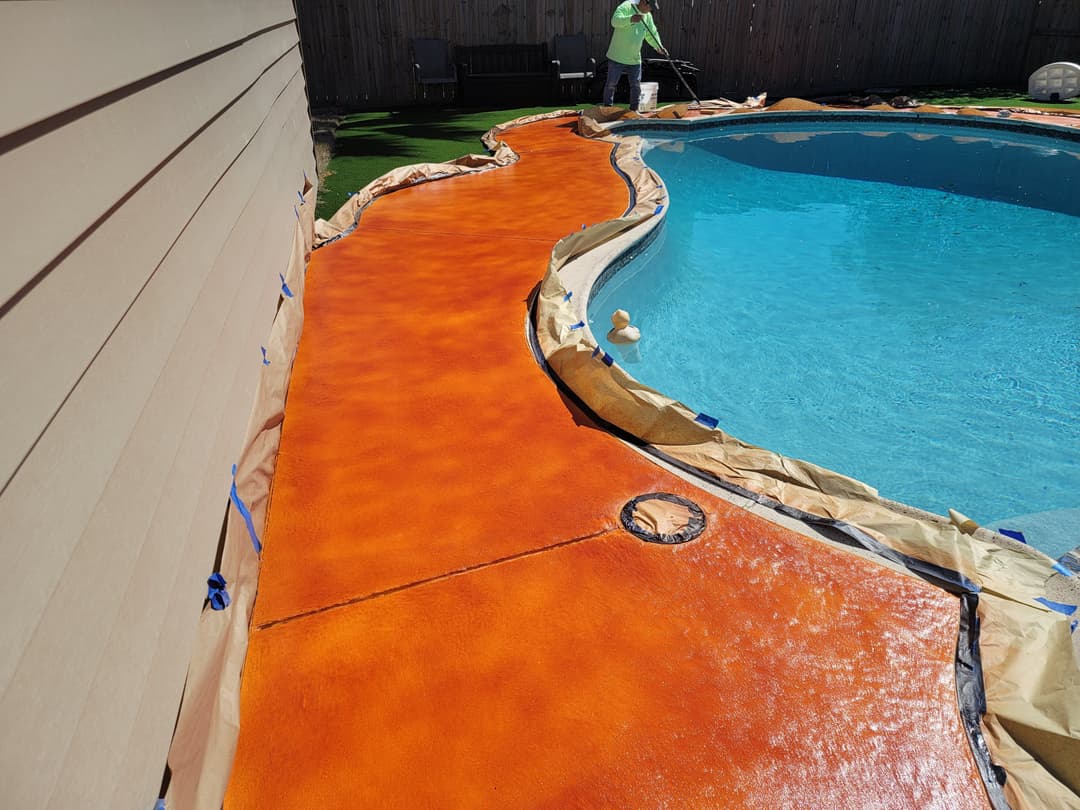 Vibrant orange pool deck under renovation next to sparkling blue water, with worker in background.