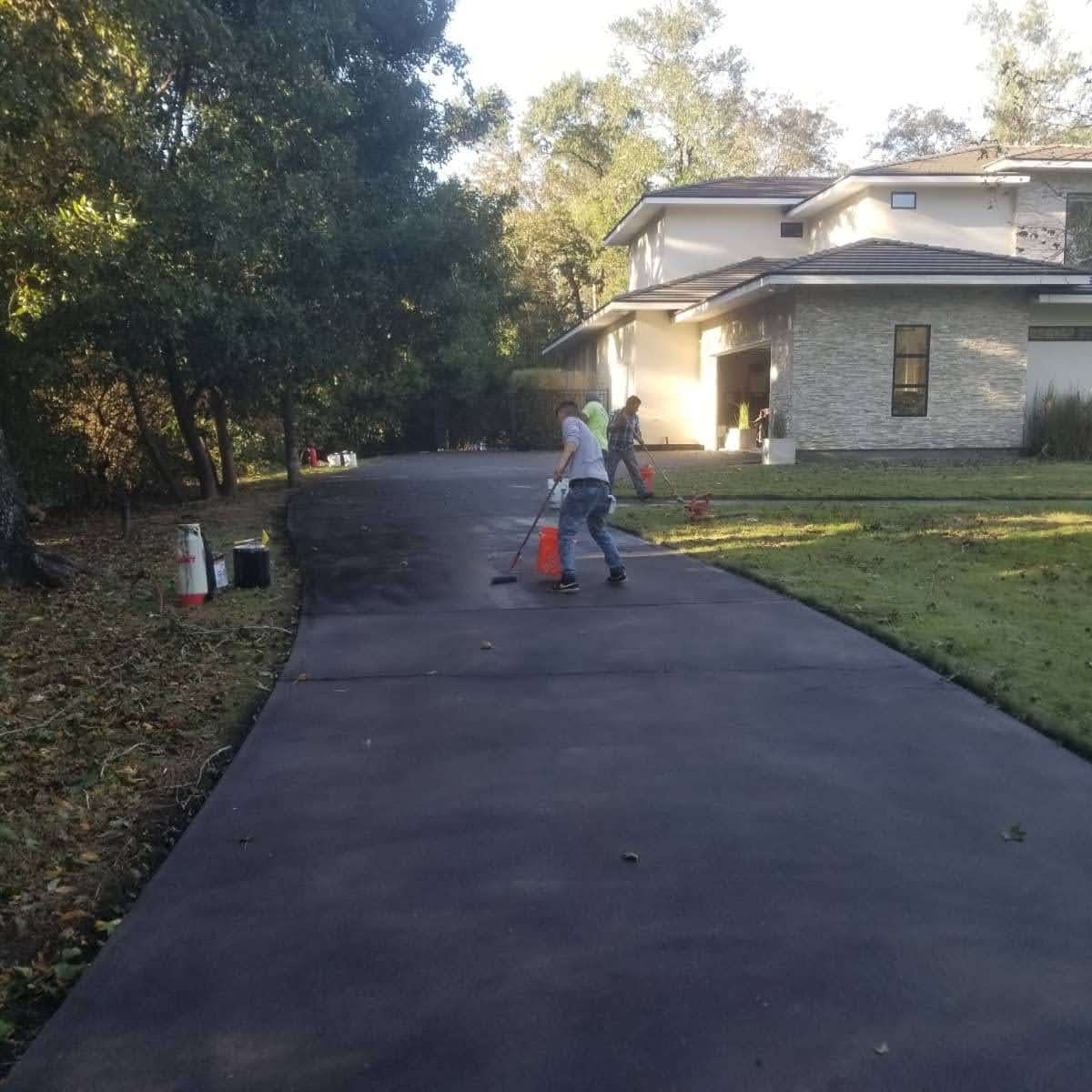 Workers applying sealant on a newly paved driveway surrounded by trees and a modern house.