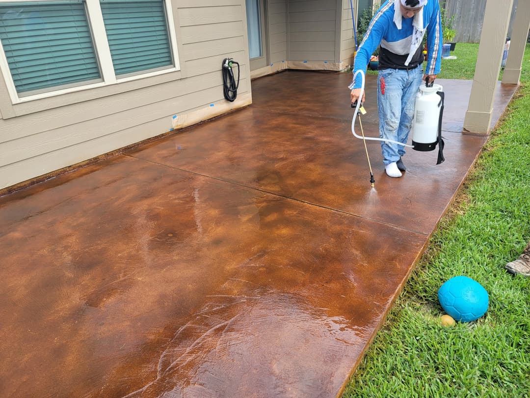 Person applying sealant to a stained concrete patio with a spray device and blue ball nearby.