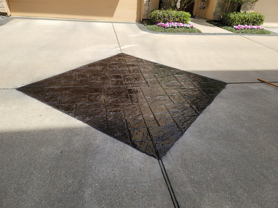Stamped concrete driveway with a dark finish and flower beds in the background.