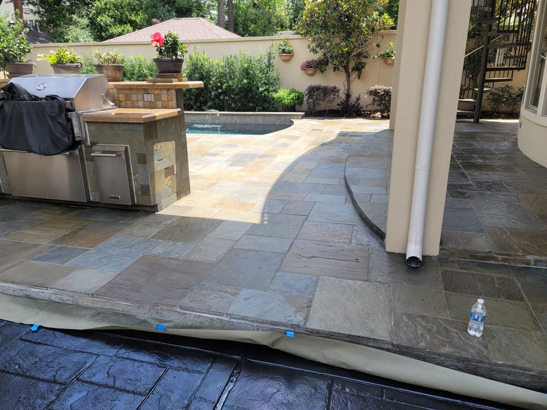 Outdoor patio with stone flooring, grill area, pool, and lush greenery in the background.