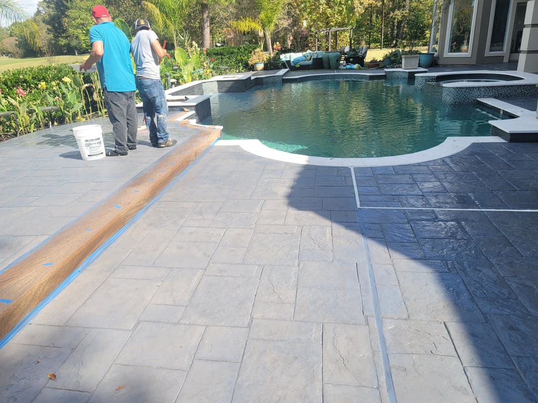 Workers applying sealant to a stamped concrete patio beside a pool in a residential backyard.