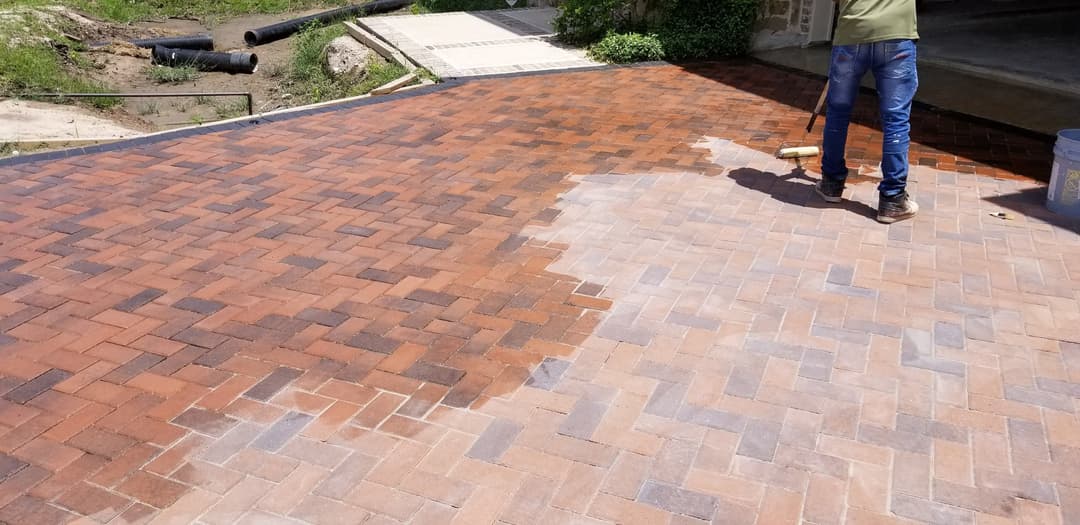 Person applying sealant to a freshly cleaned brick paver driveway in a residential setting.