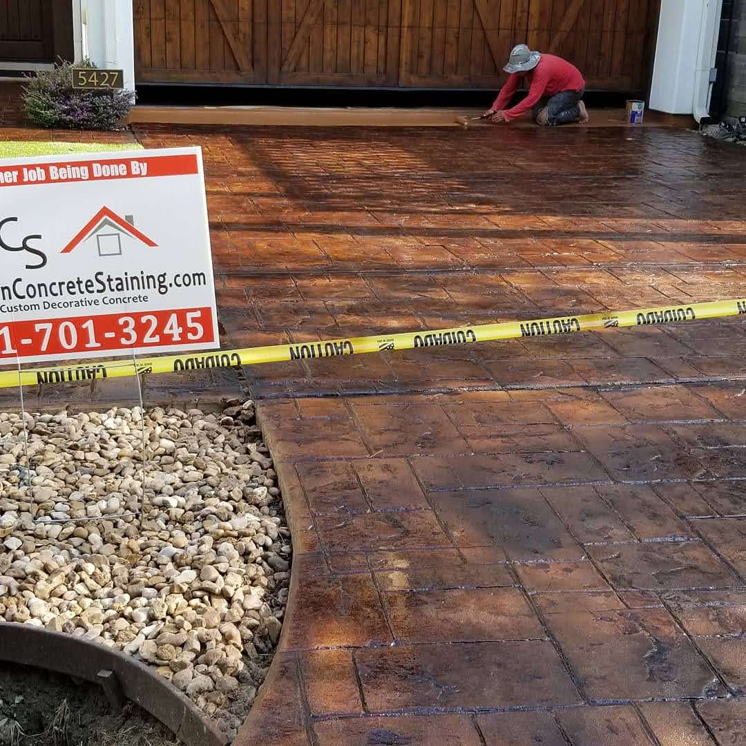 Worker applying decorative stain to a concrete driveway with caution tape and landscaping stones nearby.