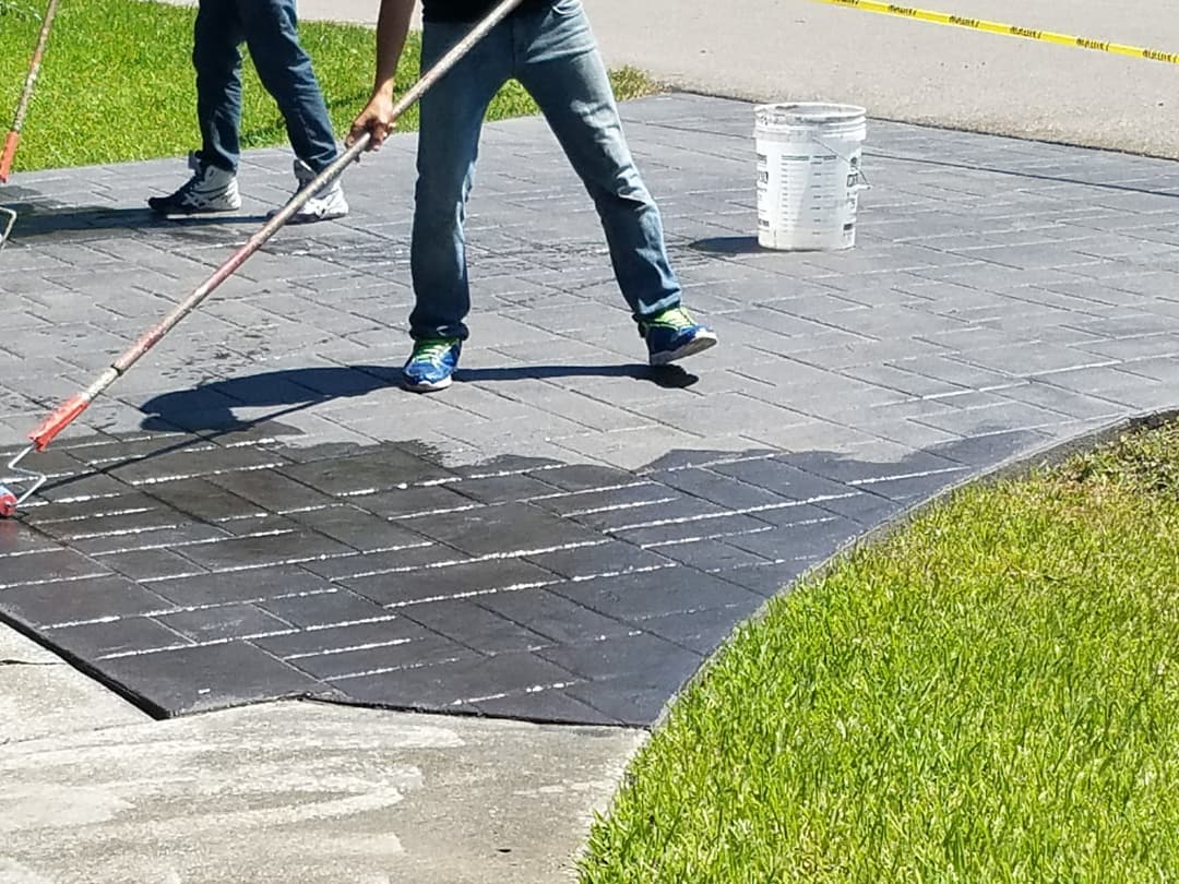 Person applying sealant to a paved driveway with a roller and bucket of sealant nearby.