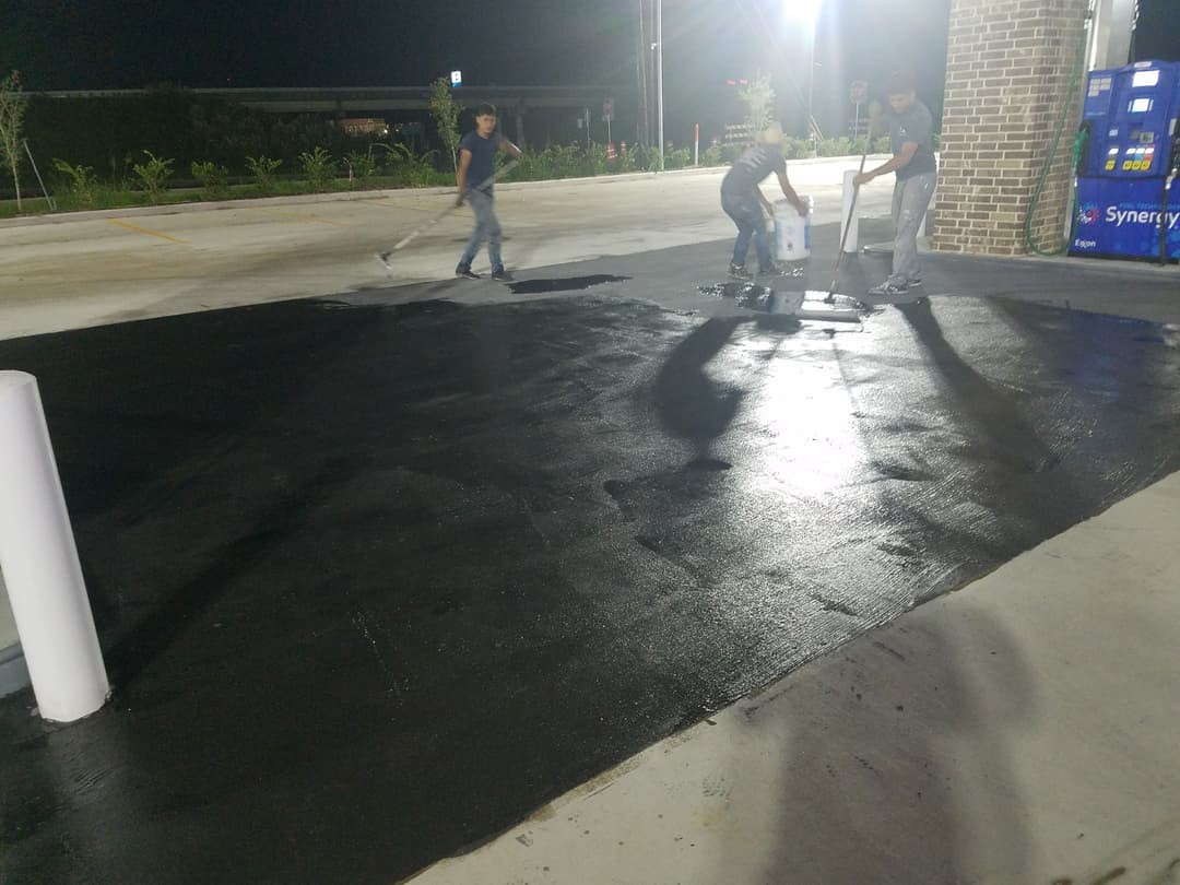 Workers applying black sealant to a parking lot surface at night.