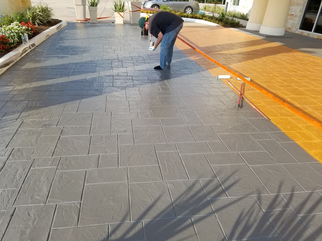 Person applying sealant on freshly stamped concrete driveway in gray and amber colors.
