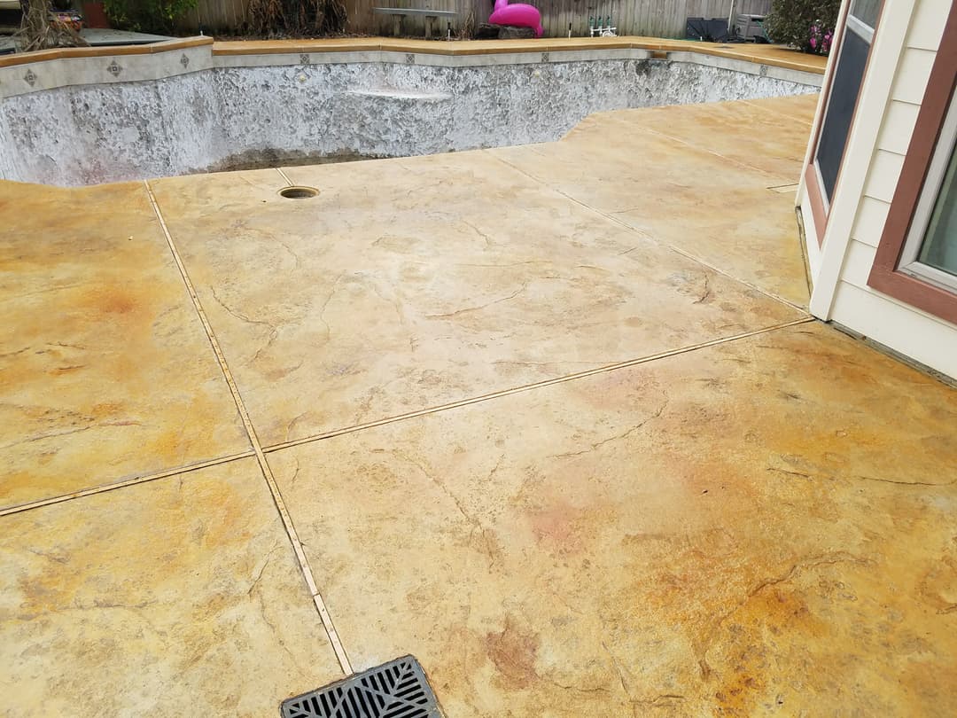 Concrete pool deck with a textured finish, near an empty swimming pool and house corner.