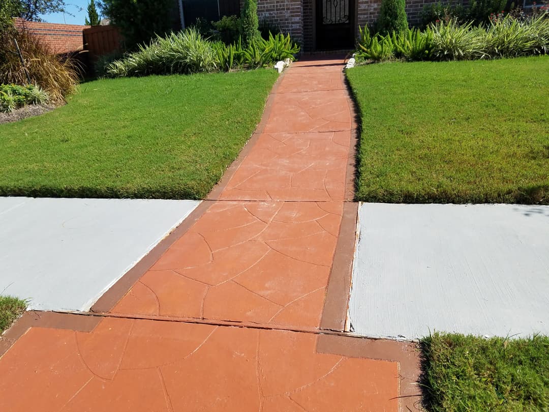 Decorative red concrete walkway leading to a house, bordered by green grass and shrubs.