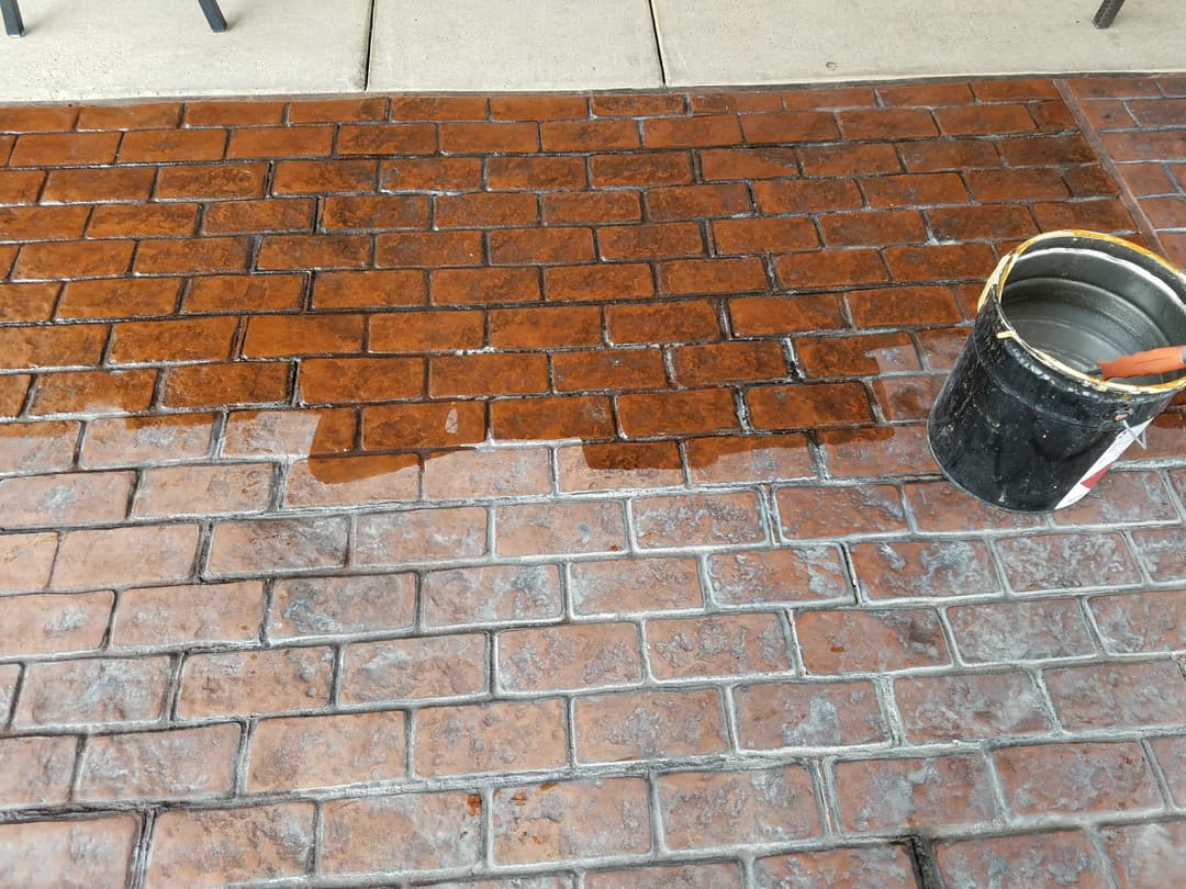 Stamped concrete patio being sealed with a black bucket on a brown brick pattern surface.
