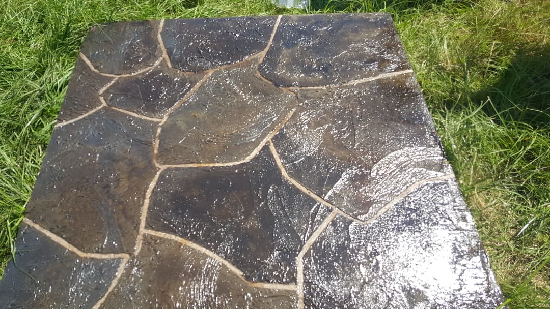 Textured concrete patio with a stone pattern, illuminated by sunlight in a grassy area.