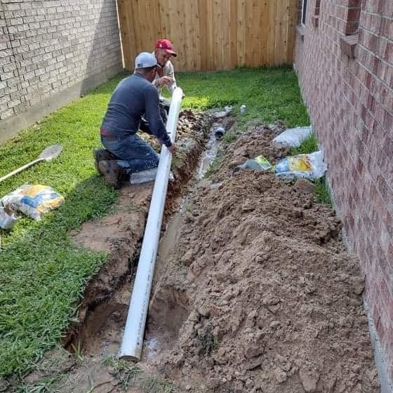 Two workers installing a drainage pipe in a residential yard with a dirt trench.