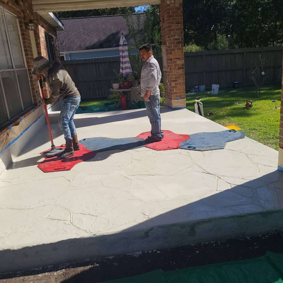 Two workers applying stamped concrete on a patio, with tools and a grassy yard in the background.