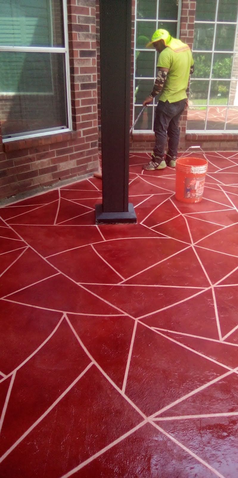 Worker applying red sealant on a patterned concrete floor in a residential patio area.