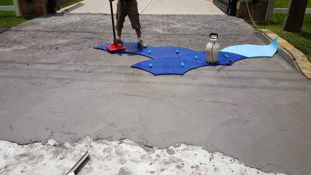 Worker applying decorative concrete overlay on a textured surface, using blue stencil and tools.