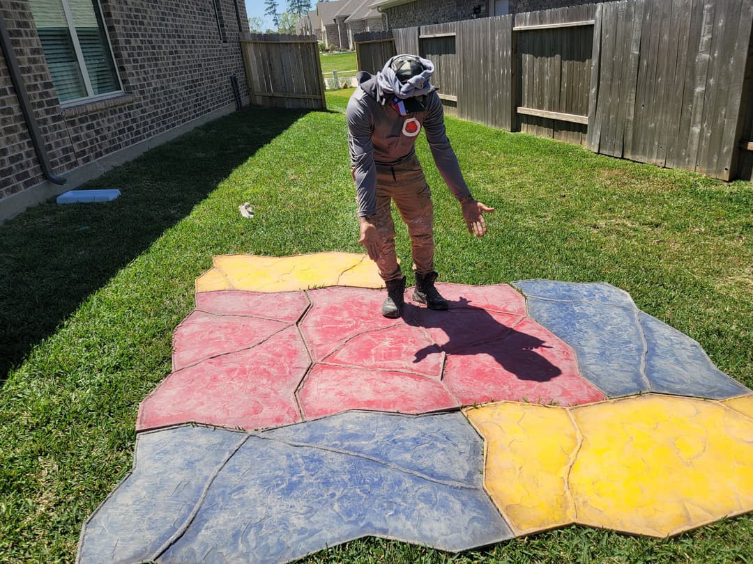 Person inspecting colorful stone-patterned outdoor mat on grass in backyard setting.