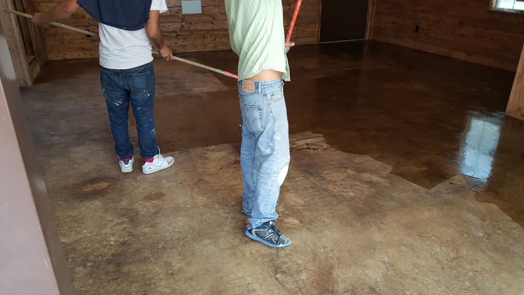 Two workers applying a glossy finish to a stained concrete floor in a wooden interior.