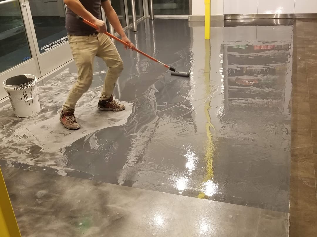 Person applying gray flooring material with a roller in a commercial space.