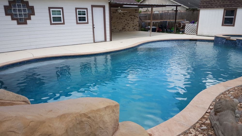 Backyard swimming pool with clear blue water surrounded by stone and a white house backdrop.